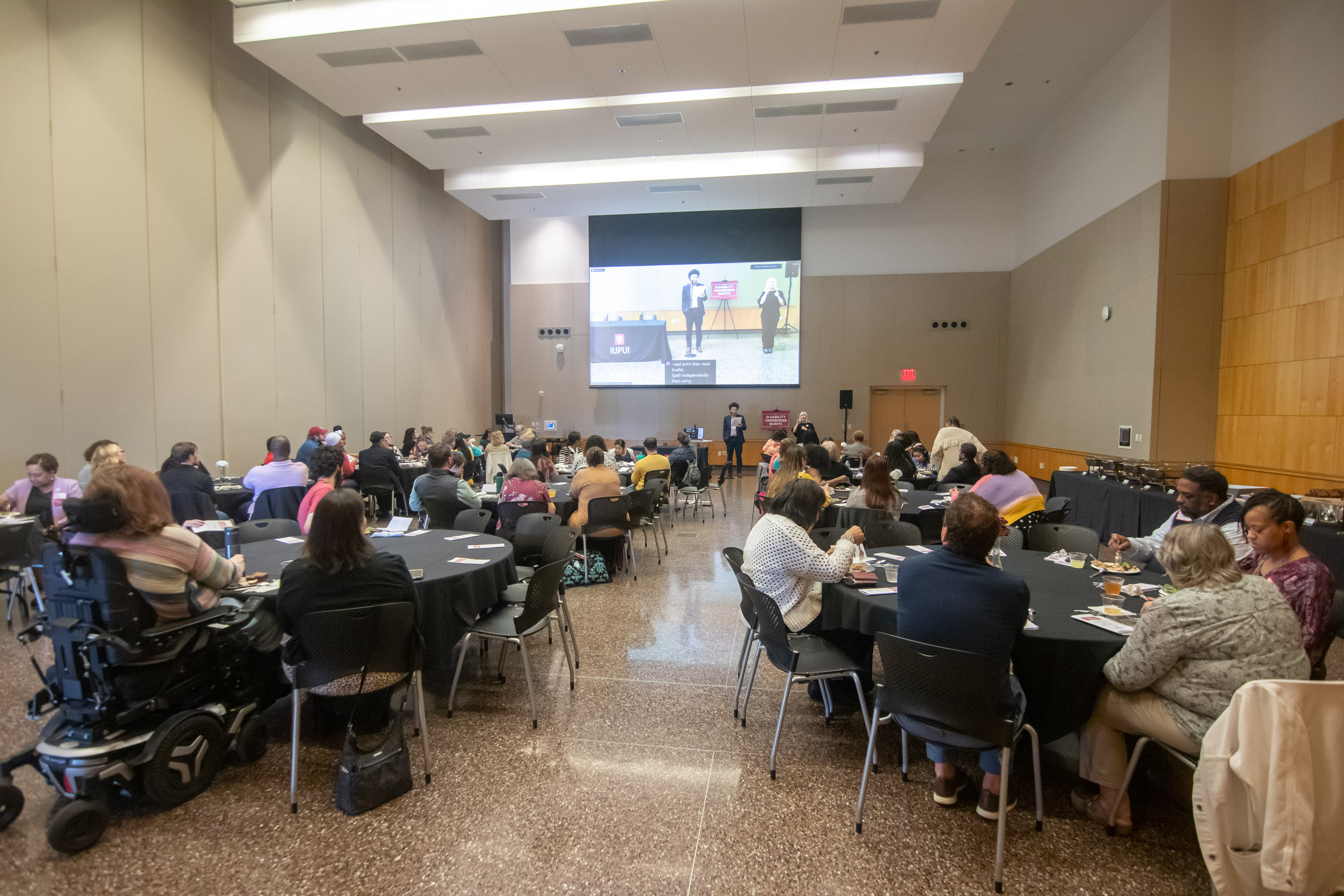 View from the back of the luncheon shows attendees finishing up lunch and Dr. Tiffany Kaiser beginning the intro with screen behind her of the recording