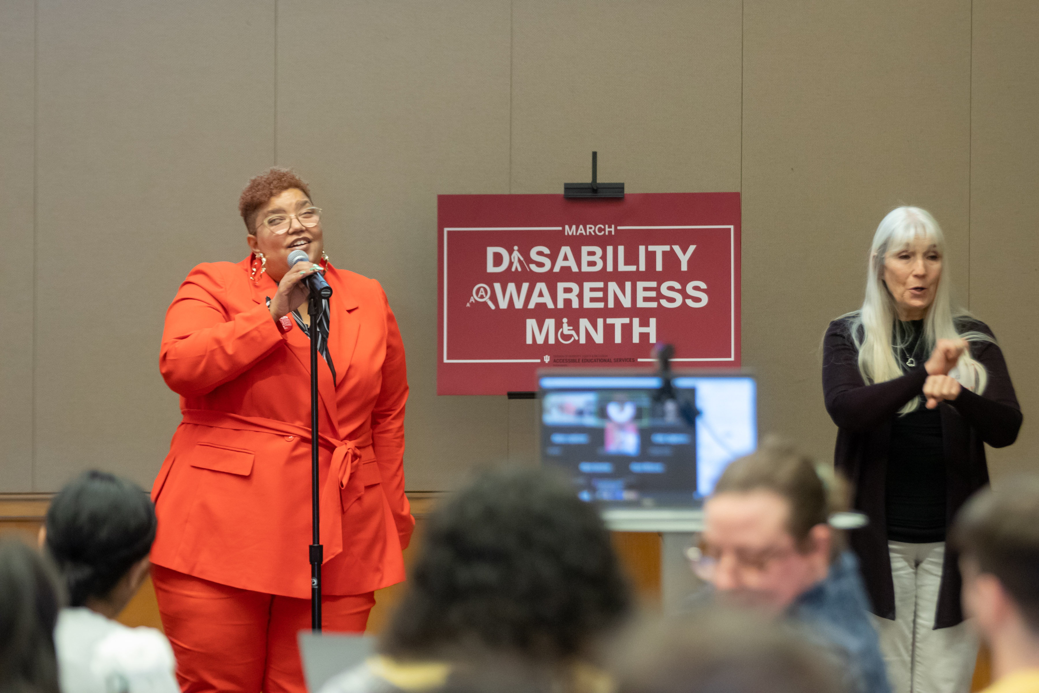 View of speaker, Dr. Katherine Betts, in her speech, with sign reading Disability Awareness Month in red, and ASL interpreter on the right