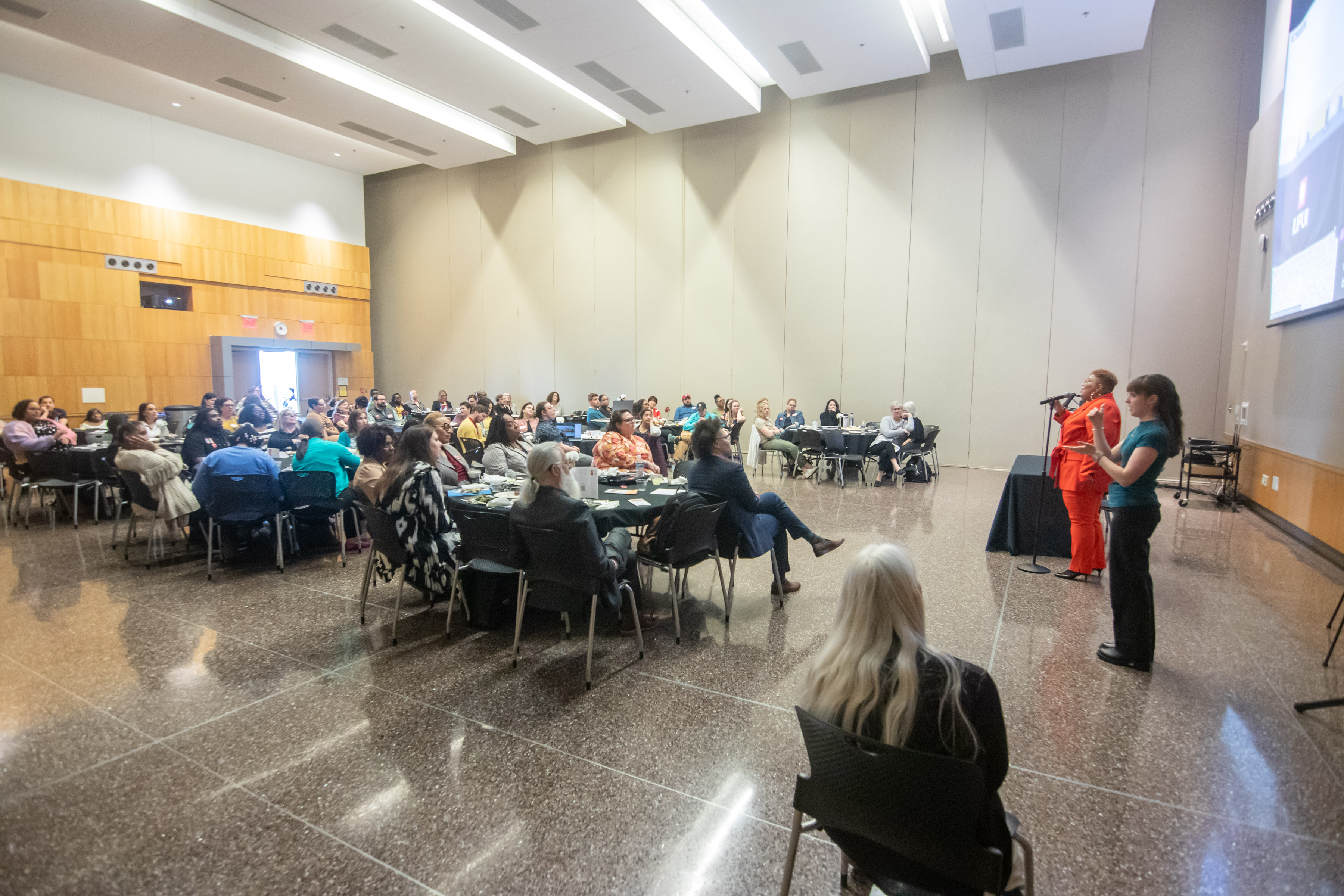 View from the side of the well attended luncheon shows the speaker standing at the microphone and an ASL interpreter standing next to her